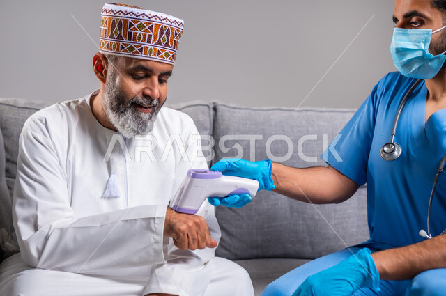 طبيب يحمل مقياس الحرارة الرقمي بالأشعة تحت الحمراء، إتباع إجراءات الوقاية و السلامة من فايروس كورونا، الوعي الصحي وتجنب إنتشار العدوى، بورتريه لممرض يرتدي اليونيفورم يقوم بفحص مريض مسن عربي خليجي عماني يرتدي الدشداشة والكمه، العمل في القطاع الطبي، خلفية رمادية