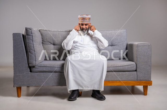 إيماءات الصداع والإحساس بالمرض, الحاجه للعلاج وطلب المساعده, بورتريه لرجل عربي خليجي عماني يرتدي الدشداشة والكمه يجلس على كنبه بريحة يمسك رأسه بيديه من شدة الألم, الشعور بالتعب والإرهاق, الإصابة بنوبات الشقيقة، صورة الجسم بالطول الكامل، خلفية رماديه