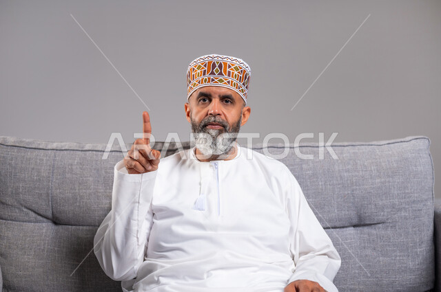 الاستفسار والاشارة إلى شيء ما, بورتريه مقرب لرجل عربي خليجي عماني يرتدي الدشداشة والكمه يجلس على كنبه بريحة يرفع اصبعه للأعلى بإيماءات التوضيح، النظرإلى الكاميرا بتعابير الاستفهام والتساؤل، خلفية رمادية