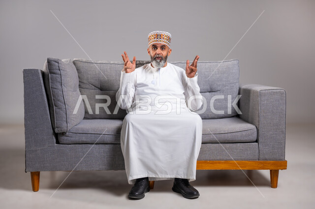 الغموض وعدم الوضوح، التساؤل والاستفهام لمعرفة موضوع مبهم، بورتريه لرجل عربي خليجي عماني يرتدي الدشداشة والكمه يجلس على كنبه بريحة يشير بيده للسؤال والاستفسار عن شيء ما، النظرالى الكاميرا بإيماءات الاستغراب، الطول الكامل للجسم, خلفية مادية