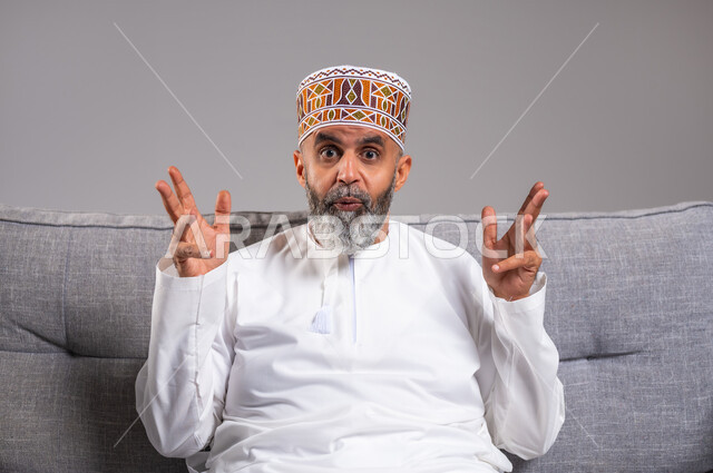 التساؤل والاستفهام لمعرفة موضوع مبهم، بورتريه مقرب لرجل عربي خليجي عماني يرتدي الدشداشة والكمه يجلس على كنبه بريحة يشير بيده للسؤال والاستفسار عن شيء ما، النظرالى الكاميرا بإيماءات الاستغراب، الغموض وعدم الوضوح، خلفية مادية