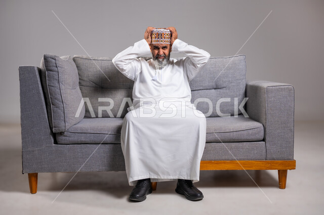 إيماءات الصداع والإحساس بالمرض, الحاجه للعلاج وطلب المساعده, بورتريه لرجل عربي خليجي عماني يرتدي الدشداشة والكمه يجلس على كنبه بريحة يمسك رأسه بيديه من شدة الألم, الشعور بالتعب والإرهاق, الإصابة بنوبات الشقيقة، صورة الجسم بالطول الكامل، خلفية رماديه