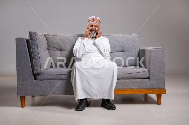 بثور منتشرة على بشرة الوجه, المشاكل الصحية والإصابة بالحساسية الجلدية, الأمراض والتهجيات وفرط التحسس, بورتريه لرجل عربي خليجي عماني يرتدي الدشداشه والكمه يضع يديه على وجهه بإيماءات الألم والحكة, الشعور بالأوجاع وعدم التحمل, الطول الكامل للجسم، خلفية رمادية