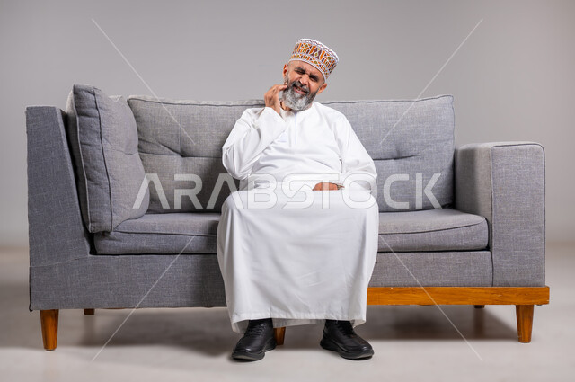 ضرورة الذهاب للطبيب والحصول على العلاج، بورتريه لرجل كبير السن عربي خليجي عماني يرتدي الدشداشه والكمه يمسك رقبته بإيماءات الألم, آلام الرقبة والعظام والعمود الفقري، إيماءات الوجه تدل على الاضطراب والتألم، الطول الكامل للجسم، خلفية رمادية