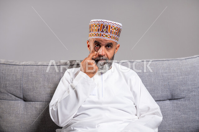 مشاكل عينيه وحالات مرضية، الحاجة للفحص والعلاج، التهاب القرنية العينية، ارتفاع ضغط العين، بورتريه مقرب لرجل مسن عربي خليجي عماني يرتدي الدشداشه والكمه يجلس على كنبه مريحة يقوم بفتح عينه باستخدام اصبع السبابة، احمرار و وجع العيون، خلفية رمادية