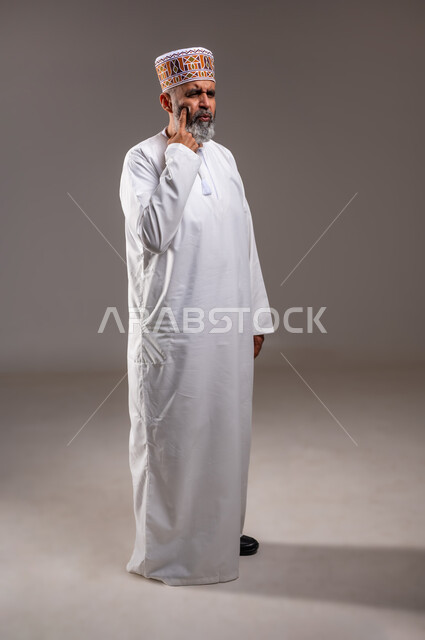 المشاكل الصحية والتهجيات والشعور بالأوجاع، بثور منتشرة على بشرة الوجه، بورتريه لرجل عربي خليجي عماني يرتدي الدشداشه والكمه يشير باصبع السبابة على مكان الإصابة، النظر الى شيء ما بايماءات الألم، صوره الجسم بالطول الكامل، خلفيه رماديه