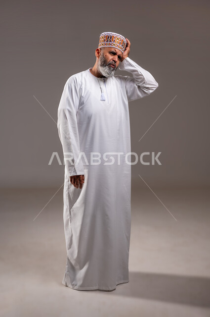 الاحساس بالشقيقية والصداع النصفي, الإرهاق وفقدان التركيز, الشعور بالصداع وألم الرأس, الاجهاد والضغط النفسي, بورتريه لرجل مسن عربي خليجي عماني يرتدي الدشداشه والكمه يضع يده على رأسه ويغمض عينيه بإيماءات الألم الشديد, الطول الكامل للجسم, خلفية رمادية