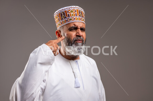 النظر الى شيء ما بايماءات الألم، المشاكل الصحية والتهجيات والشعور بالأوجاع، بثور منتشرة على بشرة الوجه، بورتريه مقرب لرجل عربي خليجي عماني يرتدي الدشداشه والكمه يشير باصبع السبابة على مكان الإصابة، خلفيه رماديه