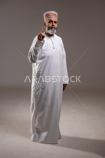 الاشارة الى شيء ما بايماءات الجدية، بورتريه  لرجل عربي خليجي عماني يرتدي الدشداشه والكمه يشير باصبع السبابة للاعلى، النظر الى الكاميرا بتعابير الغضب، حركة اصبع واشارات، صوره الجسم بالطول الكامل، خلفيه رماديه