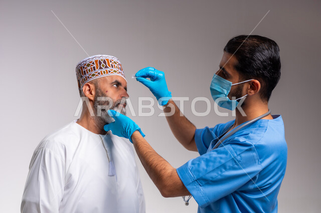 الاهتمام بالأعضاء الحساسة في الجسد, العمل في القطاع الصحي، مفهوم الطب والرعاية الصحية، بورتريه مقرب لممرض عيون مختص يرتدي اليونيفورم و والكمامه والقفازات الطبية يقوم بوضع قطرة عينية لرجل مسن عربي خليجي عماني يرتدي الدشداشة والكمه، خلفية رمادية