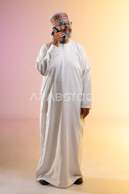 إجراء مكالمة هاتفية، ايماءات السعادة والسرور بسماع اخبار جيدة، الدردشة والتحدث مع الاصدقاء عبر الجوال، بورتريه لرجل مسن عربي خليجي عماني يرتدي الدشداشه والكمه ويضع نظارات طبية يقوم بالرد على اتصالات الجوال المحمول، الجسم بالطول الكامل، خلفية ملونة