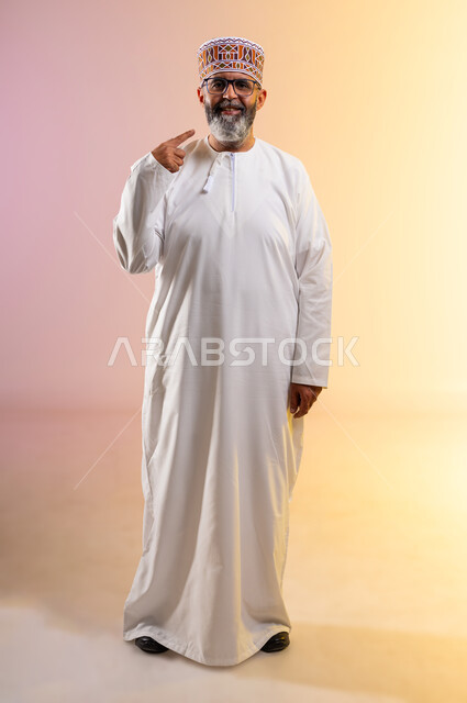 Standing straight and looking at the camera, using porcelain veneers to create a dazzling white smile, the importance of dental follow-up, care, cosmetics and whitening, portrait of an elderly Arab Gulf Omani man wearing a dishdasha and a kummah pointing to his teeth, full body image, colored background