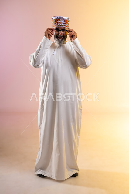 النظر إلى الكاميرا بتعابير البشاشة والسرور, الاهتمام بالأناقة والمظهر الخارجي، مفهوم طول النظر وقصره, بورتريه لرجل كبير بالسن عربي خليجي عماني يرتدي الدشداشه والكمه يمسك النظارة الطبية, الشعور بالسعادة والفرح بوضوح الرؤية, الطول الكامل للجسم، خلفية ملونة