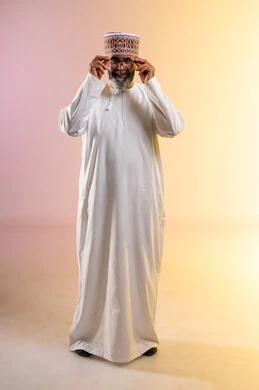 Looking at the camera with expressions of joy and happiness, interest in elegance and appearance, concept of farsightedness and nearsightedness, portrait of an elderly Arab Gulf Omani man wearing a dishdasha and a sleeve holding eyeglasses, feeling happy and joyful with clear vision, full body length, colorful background