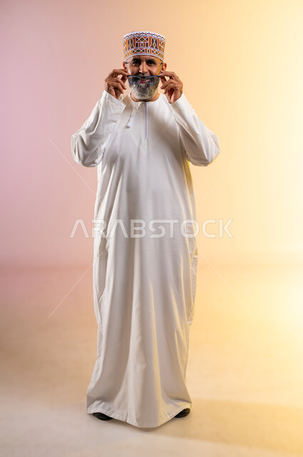 مفهوم طول النظر وقصره, بورتريه لرجل كبير بالسن عربي خليجي عماني يرتدي الدشداشه والكمه يضع النظارة الطبية بإيماءات السرور, النظر إلى الكاميرا بتعابير التمعن والتدقيق, صورة الجسم بالطول الكامل، الاهتمام بالأناقة والمظهر الخارجي، خلفية ملونة