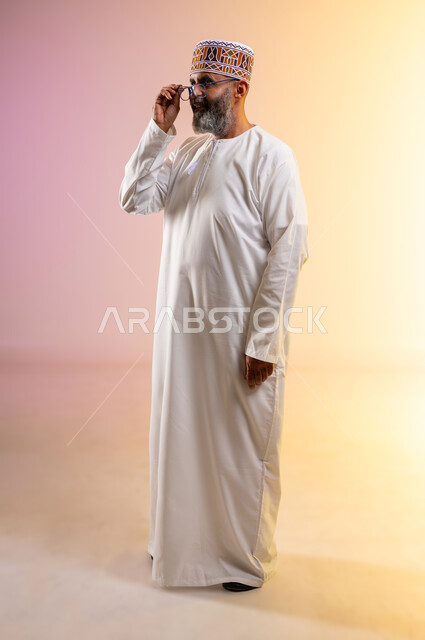 Concern for elegance and appearance, concept of farsightedness and nearsightedness, portrait of an elderly Arab Gulf Omani man wearing a dishdasha and a kilt holding glasses with gestures of pleasure, contemplating and examining something, full-length body image, colored background