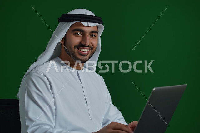 إستخدام اجهزة تقنية حديثة، بورتريه مقرب لرجل أعمال عربي خليجي سعودي مبتسم يرتدي الزي السعودي يقوم بانجاز مهام العمل عبر جهاز الحاسب المحمول، مفهوم إدارة المشاريع عن بعد، النظر الى الكاميرا بايماءات السرور والسعادة، خلفية خضراء 