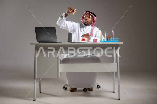 البحث وإجراء التفاعلات الكيميائية, اكتساب المعرفة والخبرة بإيماءات الدقة والتركيز، الدراسات والتجارب في بيئة عملية متطورة, بورتريه لرجل عربي خليجي سعودي يرتدي الزي التقليدي و نظارة الوقاية يجلس امام الحاسوب المحمول يقوم بتجربة علمية, صورة لكامل الجسم، خلفية رمادية