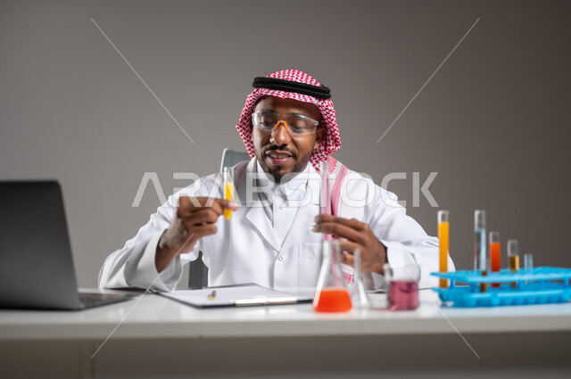 Gaining knowledge and experience with gestures of precision and focus, studies and experiments in an advanced practical environment, development, research and conducting chemical reactions, close-up portrait of a Saudi Arabian Gulf man wearing traditional dress and goggles sitting in front of a laptop doing a scientific experiment, gray background