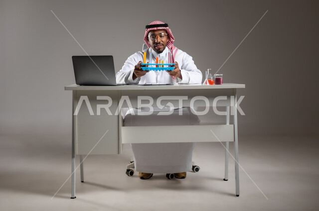إيماءات الدقة والتركيز، إجراء التفاعلات الكيميائية, اكتساب المعرفة والخبرة، الدراسات والتجارب في بيئة عملية متطورة, بورتريه لرجل عربي خليجي سعودي يرتدي الزي التقليدي و نظارة الوقاية يجلس امام الحاسوب المحمول يقوم بتجربة علمية, صورة لكامل الجسم، خلفية رمادية