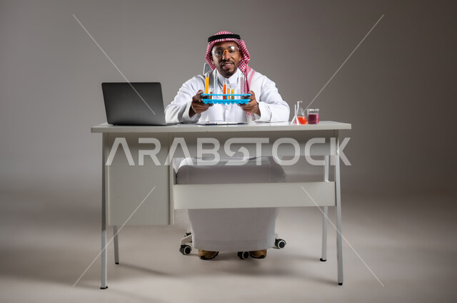 البحث وإجراء التفاعلات الكيميائية, اكتساب المعرفة والخبرة بإيماءات الدقة والتركيز، الدراسات والتجارب في بيئة عملية متطورة, بورتريه لرجل عربي خليجي سعودي يرتدي الزي التقليدي و نظارة الوقاية يجلس امام الحاسوب المحمول يقوم بتجربة علمية, صورة لكامل الجسم، خلفية رمادية