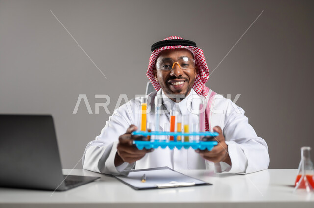 اكتساب المعرفة والخبرة بإيماءات الدقة والتركيز، الدراسات والتجارب في بيئة عملية متطورة, التطوير والبحث وإجراء التفاعلات الكيميائية, بورتريه مقرب لرجل عربي خليجي سعودي يرتدي الزي التقليدي و نظارة الوقاية يجلس امام الحاسوب المحمول يقوم بتجربة علمية, خلفية رمادية