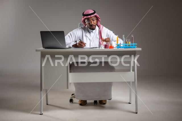 مفهوم تدوين الملاحظات، إجراء التفاعلات الكيميائية, اكتساب المعرفة والخبرة، الدراسات والتجارب في بيئة عملية متطورة, بورتريه لرجل عربي خليجي سعودي يرتدي الزي التقليدي و نظارة الوقاية يجلس امام الحاسوب المحمول يقوم بتجربة علمية, صورة لكامل الجسم، خلفية رمادية