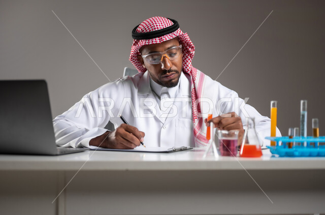 إجراء التفاعلات الكيميائية, مفهوم تدوين الملاحظات، اكتساب المعرفة والخبرة، الدراسات والتجارب في بيئة عملية متطورة, بورتريه مقرب لرجل عربي خليجي سعودي يرتدي الزي التقليدي و نظارة الوقاية يجلس امام الحاسوب المحمول يقوم بتجربة علمية, خلفية رمادية