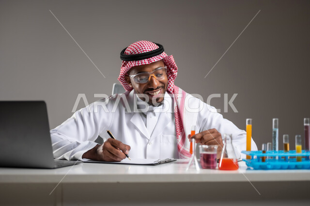 مفهوم تدوين الملاحظات، إجراء التفاعلات الكيميائية, اكتساب المعرفة والخبرة، الدراسات والتجارب في بيئة عملية متطورة, بورتريه مقرب لرجل عربي خليجي سعودي يرتدي الزي التقليدي و نظارة الوقاية يجلس امام الحاسوب المحمول يقوم بتجربة علمية, خلفية رمادية
