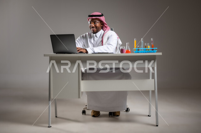 القيام بالتجارب في بيئة عملية متطورة, استخدام الاجهزة التقنية، إجراء التفاعلات الكيميائية، بورتريه مقرب لايدي رجل عربي خليجي سعودي يرتدي الزي التقليدي و نظارة الوقاية يجلس امام الحاسوب المحمول يقوم بالعمل على الحاسوب المحمول, صورة لكامل الجسم، خلفية رمادية