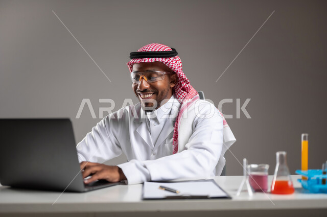 استخدام الاجهزة التقنية، إجراء التفاعلات الكيميائية, اكتساب المعرفة والخبرة، الدراسات والتجارب في بيئة عملية متطورة, بورتريه مقرب لايدي رجل عربي خليجي سعودي يرتدي الزي التقليدي و نظارة الوقاية يجلس امام الحاسوب المحمول يقوم بالعمل على الحاسوب المحمول, خلفية رمادية