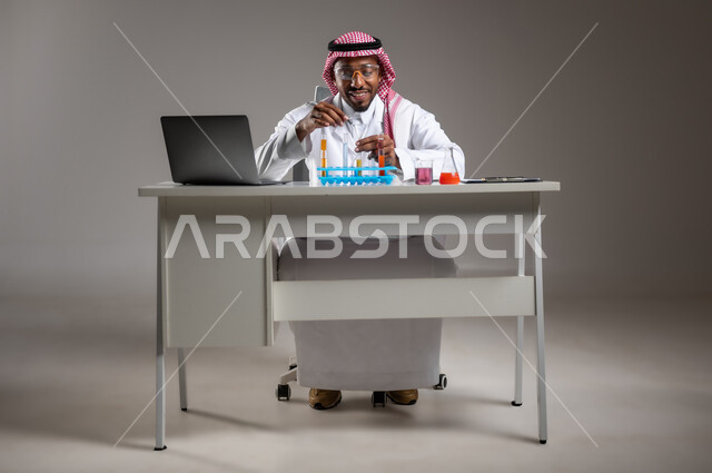 البحث وإجراء التفاعلات الكيميائية, اكتساب المعرفة والخبرة بإيماءات الدقة والتركيز، الدراسات والتجارب في بيئة عملية متطورة, بورتريه لرجل عربي خليجي سعودي يرتدي الزي التقليدي و نظارة الوقاية يجلس امام الحاسوب المحمول يقوم بتجربة علمية, صورة لكامل الجسم، خلفية رمادية
