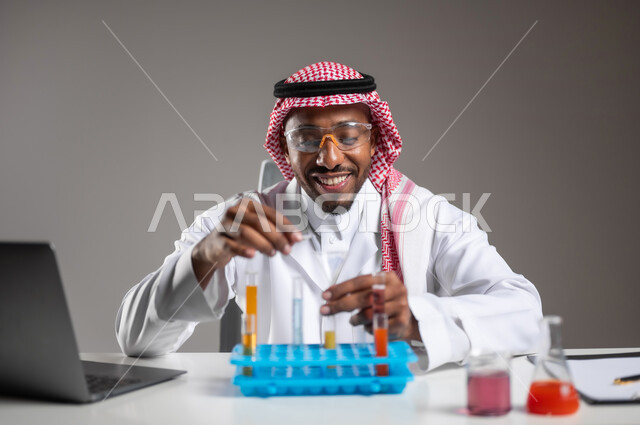 Gaining knowledge and experience with gestures of precision and focus, studies and experiments in an advanced practical environment, development, research and conducting chemical reactions, close-up portrait of a Saudi Arabian Gulf man wearing traditional dress and goggles sitting in front of a laptop doing a scientific experiment, gray background