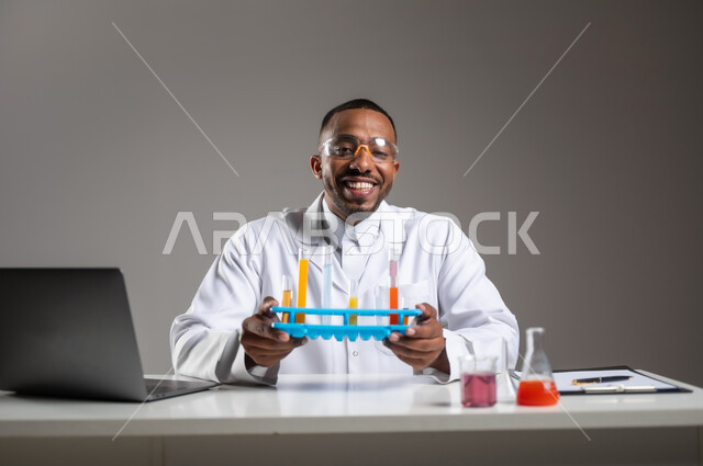 التطوير والبحث وإجراء التفاعلات الكيميائية, اكتساب المعرفة والخبرة بإيماءات الدقة والتركيز، الدراسات والتجارب في بيئة عملية متطورة, بورتريه مقرب لرجل عربي خليجي سعودي يرتدي الزي التقليدي و نظارة الوقاية يجلس امام الحاسوب المحمول يقوم بتجربة علمية, خلفية رمادية