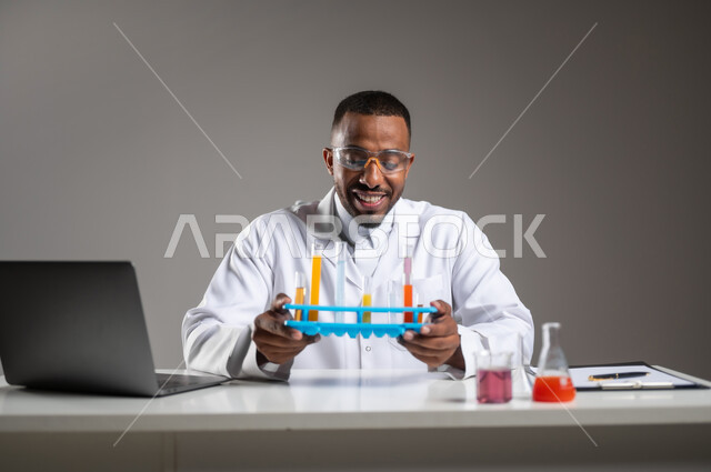 Gaining knowledge and experience with gestures of precision and focus, studies and experiments in an advanced practical environment, development, research and conducting chemical reactions, close-up portrait of a Saudi Arabian Gulf man wearing traditional dress and goggles sitting in front of a laptop doing a scientific experiment, gray background