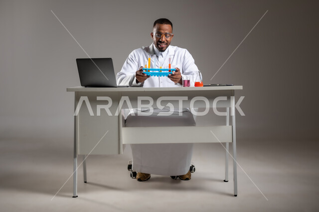 إيماءات الدقة والتركيز، إجراء التفاعلات الكيميائية, اكتساب المعرفة والخبرة، الدراسات والتجارب في بيئة عملية متطورة, بورتريه لرجل عربي خليجي سعودي يرتدي الزي التقليدي و نظارة الوقاية يجلس امام الحاسوب المحمول يقوم بتجربة علمية, صورة لكامل الجسم، خلفية رمادية