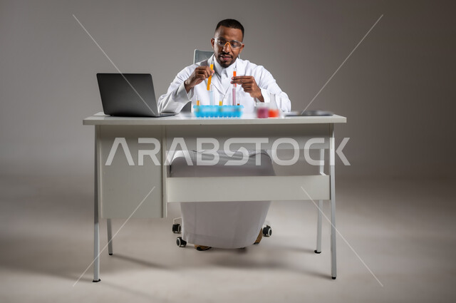 البحث وإجراء التفاعلات الكيميائية, اكتساب المعرفة والخبرة بإيماءات الدقة والتركيز، الدراسات والتجارب في بيئة عملية متطورة, بورتريه لرجل عربي خليجي سعودي يرتدي الزي التقليدي و نظارة الوقاية يجلس امام الحاسوب المحمول يقوم بتجربة علمية, صورة لكامل الجسم، خلفية رمادية