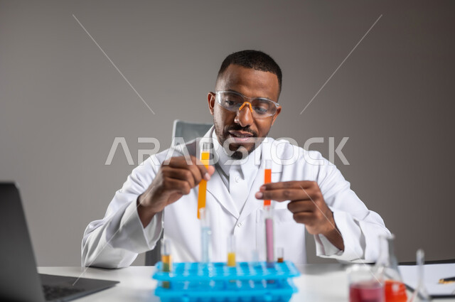 التطوير والبحث وإجراء التفاعلات الكيميائية, اكتساب المعرفة والخبرة بإيماءات الدقة والتركيز، الدراسات والتجارب في بيئة عملية متطورة, بورتريه مقرب لرجل عربي خليجي سعودي يرتدي الزي التقليدي و نظارة الوقاية يجلس امام الحاسوب المحمول يقوم بتجربة علمية, خلفية رمادية