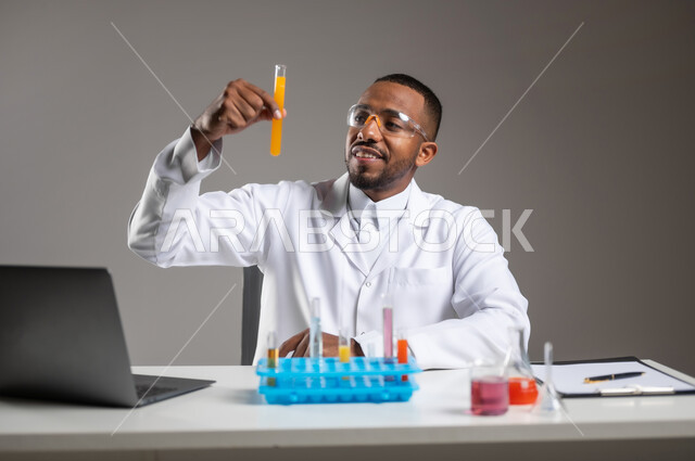 اكتساب المعرفة والخبرة بإيماءات الدقة والتركيز، الدراسات والتجارب في بيئة عملية متطورة, التطوير والبحث وإجراء التفاعلات الكيميائية, بورتريه مقرب لرجل عربي خليجي سعودي يرتدي الزي التقليدي و نظارة الوقاية يجلس امام الحاسوب المحمول يقوم بتجربة علمية, خلفية رمادية