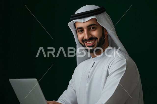 Integrating work with technology and technology, a close-up picture of a smiling Saudi Gulf Arab man wearing a casual outfit working on a laptop , office administrative professions and jobs, the concept of managing and organizing business affairs through technical devices, looking at the camera with happy gestures