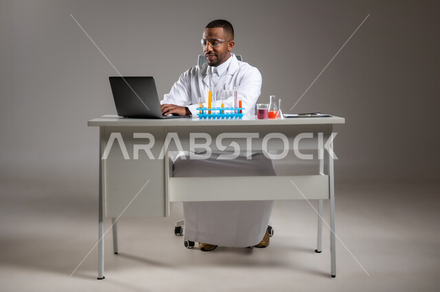 Conducting research and interactions in educational laboratories, using modern technologies, conducting experiments in a chemistry lab, portrait of a Saudi Arabian Gulf man wearing traditional dress and coat, wearing safety glasses, taking notes on a laptop, full body image, gray background