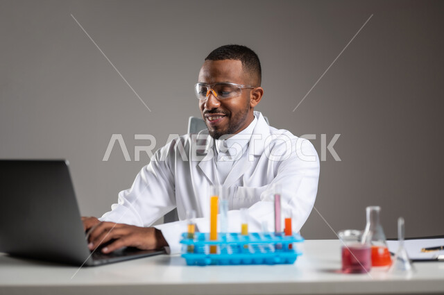 استخدام التقنيات الحديثة، القيام بالبحوث والتفاعلات في المختبرات التعليمية، إجراء التجارب العلمية في معمل الكيمياء، بورتريه مقرب لرجل عربي خليجي سعودي يرتدي الزي التقليدي والمعطف يضع نظارة الحماية يقوم بتدوين الملاحظات على جهاز الحاسوب المحمول، خلفية رمادية