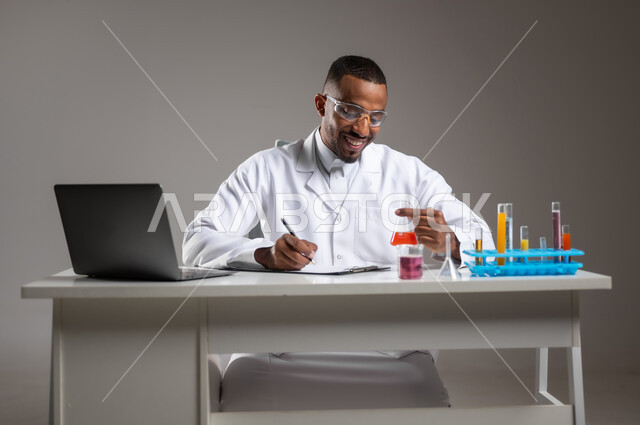 إجراء تجارب علمية في معمل الكيمياء، بورتريه مقرب لرجل عربي خليجي سعودي يرتدي الزي التقليدي والمعطف الأبيض يضع نظارة الحماية يحمل بيده أنابيب الاختبار, القيام بالبحوث والتفاعلات في المختبرات التعليمية، مفهوم تدوين الملاحظات، خلفية رمادية