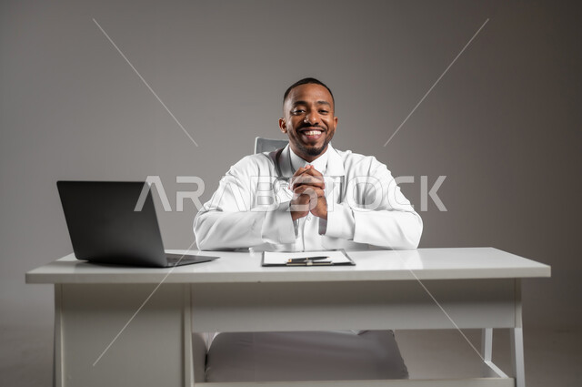 النظر إلى الكاميرا بايماءات الثقة بالنفس، العمل في القطاع الطبي، اجراء الاستشارات الطبيه، استخدام التكنولوجيا في البحث والتطوير، تقديم الخدمات للمرضى، بورتريه مقرب لطبيب عربي خليجي سعودي يرتدي الثوب التقليدي والمعطف الابيض يجلس بايدي متقاطعة، خلفيه رماديه