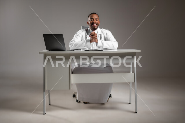 العمل في القطاع الطبي، اجراء الاستشارات الطبيه، استخدام التكنولوجيا في البحث والتطوير، تقديم الخدمات للمرضى بكل كفاءه، بورتريه لطبيب عربي خليجي سعودي يرتدي الثوب التقليدي والمعطف الابيض ينظر إلى الكاميرا بايماءات الثقة بالنفس، صورة لكامل الجسم، خلفيه رماديه