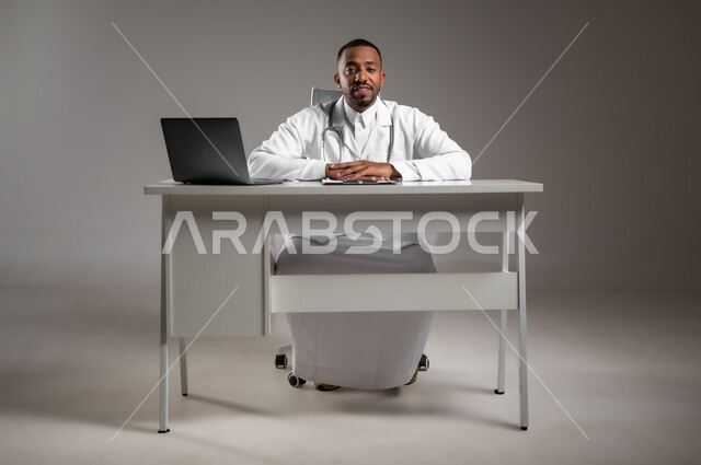 النظر إلى الكاميرا بايماءات الثقة بالنفس، العمل في القطاع الطبي، اجراء الاستشارات الطبيه، استخدام التكنولوجيا في البحث والتطوير، تقديم الخدمات للمرضى بكل كفاءه، بورتريه لطبيب عربي خليجي سعودي يرتدي الثوب التقليدي والمعطف الابيض، صورة لكامل الجسم، خلفيه رماديه