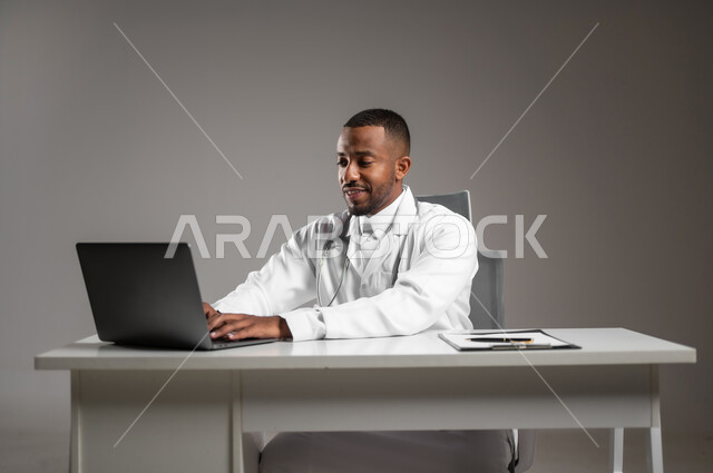 تقديم الخدمات للمرضى بكل كفاءه، اجراء الاستشارات الطبيه عن بعد، استخدام التكنولوجيا في البحث والتطوير، بورتريه مقرب لطبيب عربي خليجي سعودي يرتدي الثوب التقليدي والمعطف الابيض يعمل على جهاز الحاسوب المحمول، مهن ووظائف صحية، خلفيه رماديه