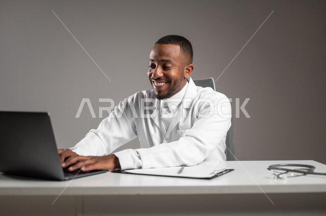 تقديم الخدمات للمرضى بكل كفاءه، اجراء الاستشارات الطبيه عن بعد، استخدام التكنولوجيا في البحث والتطوير، بورتريه مقرب لطبيب عربي خليجي سعودي يرتدي الثوب التقليدي والمعطف الابيض يعمل على جهاز الحاسوب المحمول، مهن ووظائف صحية، خلفيه رماديه