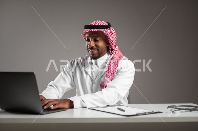 استخدام التكنولوجيا في البحث والتطوير، تقديم الخدمات للمرضى بكل كفاءه، اجراء الاستشارات الطبيه عن بعد، بورتريه مقرب لطبيب عربي خليجي سعودي يرتدي الثوب التقليدي والمعطف الابيض يعمل على جهاز الحاسوب المحمول، مهن ووظائف صحية، خلفيه رماديه