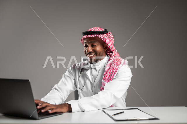 Providing services to patients efficiently, conducting remote medical consultations, using technology in research and development, close-up portrait of a Saudi Arabian Gulf doctor wearing traditional thobe and white coat working on laptop, health professions and jobs, gray background